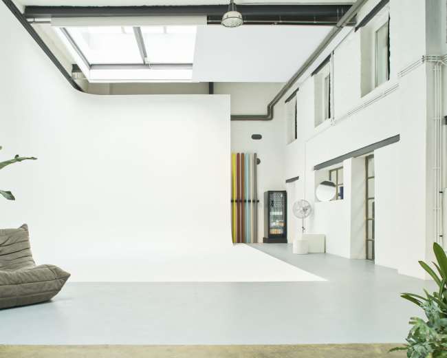 The image shows a spacious studio with a white backdrop, a large window allowing natural light, a fan, a couch, and colorful vertical stripes on one wall.