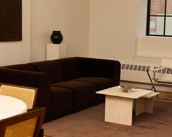 The image shows a modern living room featuring a dark sofa, a small concrete coffee table, and a chair beside a large window.