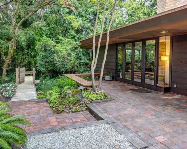 The image shows a modern house with a patio made of brick pavers, surrounded by lush greenery and landscaped areas.