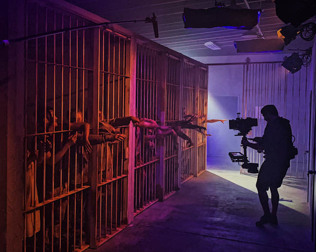 A film crew captures a scene in a dimly lit corridor of a prison set, with actors reaching through bars in the background while a camera operator films.