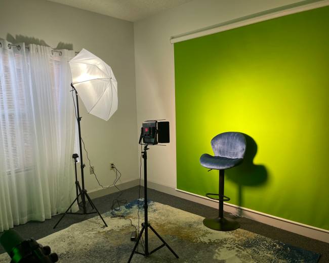 A minimal studio setup with a green backdrop, a blue chair, a lighting umbrella, and a spotlight aimed at the wall.