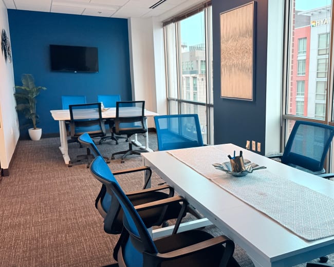 A conference room features a large white table surrounded by blue chairs, with a window offering a view of a building outside.