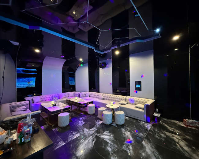 The image shows a modern, spacious lounge area with white sofas, small tables, and a sleek black and marble floor, illuminated by colorful lights.
