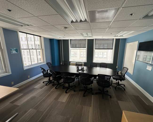 A conference room features a large, oval table surrounded by ten black chairs, with large windows letting in natural light and a wall-mounted television.