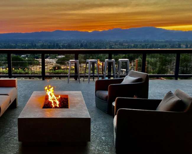 A patio area features modern seating around a central fire pit with a view of the sunset over distant mountains.