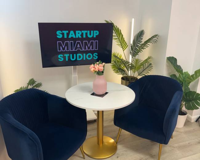 The image shows a small modern café-like setup with two navy blue chairs and a round white table, accompanied by a television displaying "STARTUP MIAMI STUDIOS," and surrounded by potted plants.