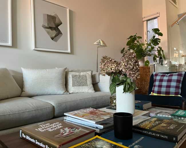 A light-colored sofa is positioned in a living room with framed art on the wall, a variety of books stacked on a coffee table, a vase of flowers, and a potted plant in the background.