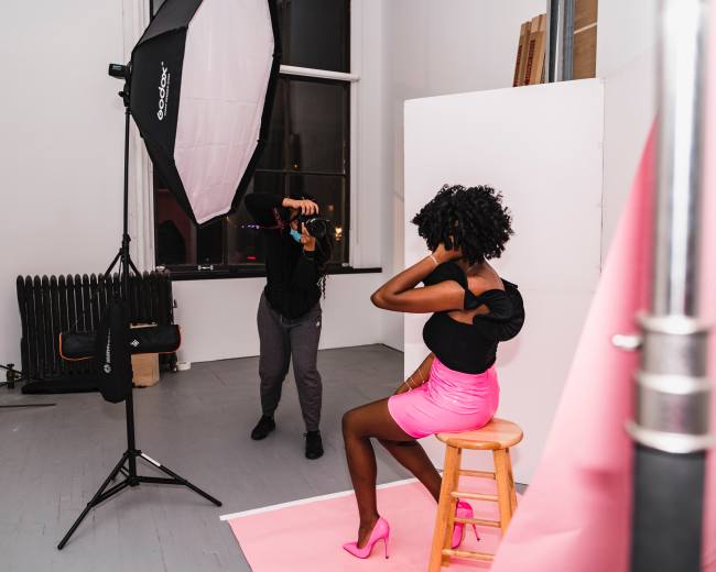 A photographer captures a model in a studio setting, featuring a pink backdrop and professional lighting equipment.