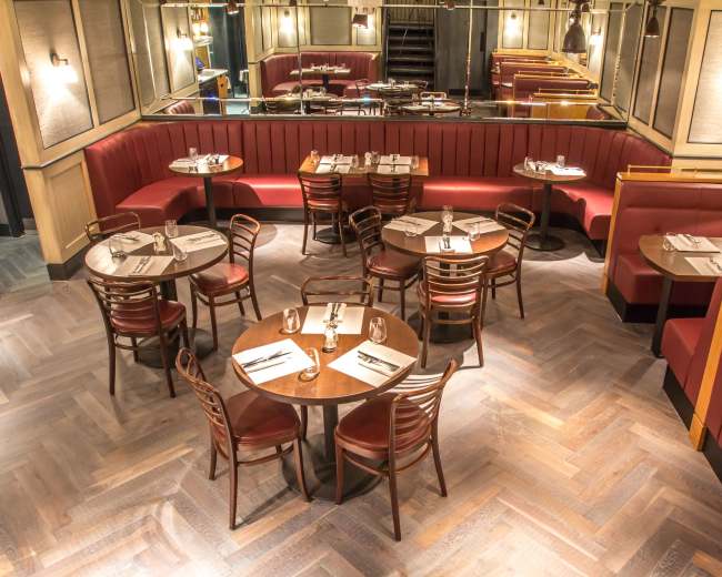 The image shows a restaurant interior featuring wooden tables surrounded by chairs and banquettes, with a mirrored wall reflecting the seating arrangement.