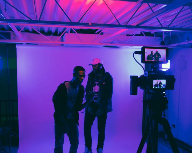 Two men are posing in front of a camera with a white backdrop and colorful lighting.