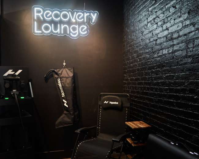 The image shows a small recovery lounge with a neon sign that reads "Recovery Lounge," a treatment chair, and equipment arranged against a black brick wall.