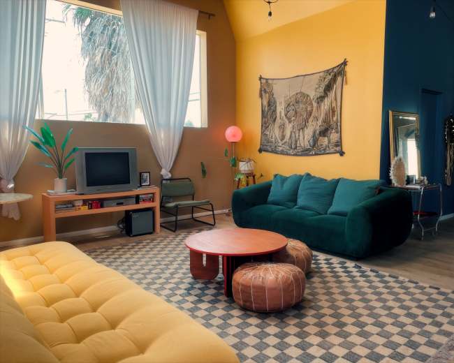 The image shows a brightly colored living room with yellow walls, a green couch, a large yellow sofa, a round coffee table, and decorative elements like plants and wall art.