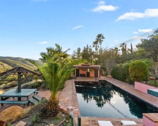 The image shows a rectangular swimming pool surrounded by palm trees, a gazebo, and a wooden deck, set against a backdrop of hills and clear blue skies.