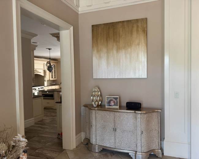 A console table with a decorative lamp and a framed photograph is positioned against a wall featuring a gold-gradient artwork, leading to an open kitchen in the background.