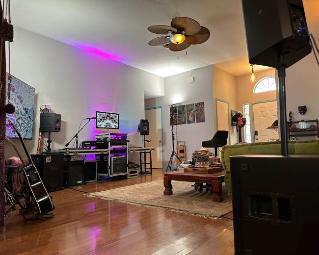 The image shows a spacious room featuring a music setup with speakers, a guitar, and colorful lighting, along with a wooden coffee table and doorways leading to other areas.