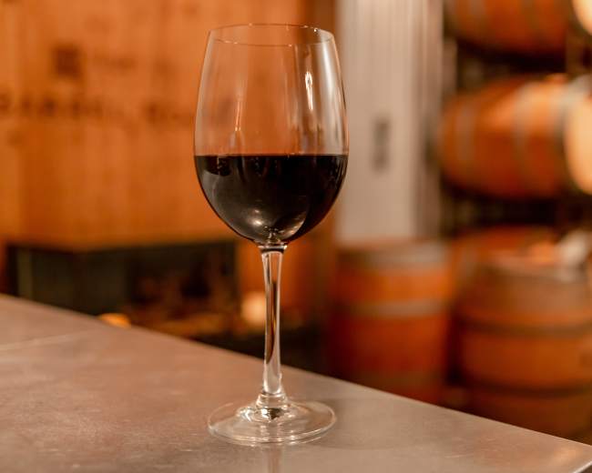 A glass of red wine is placed on a metallic surface with wooden barrels visible in the background.