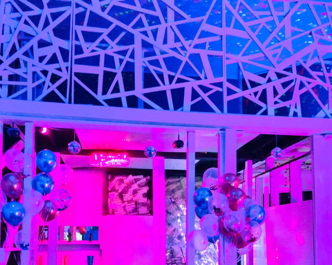 A modern lounge area with a curved sofa, a round table, and colorful balloons arranged near the columns, all illuminated by purple and blue lighting.