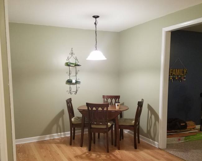 A small dining area features a wooden table with four chairs, illuminated by a pendant light, and a green wall with a decorative shelf.