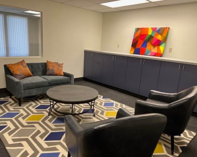 The image shows a modern office space with a gray sofa, two black chairs, a round coffee table, and colorful artwork on the wall, all set on a patterned rug.