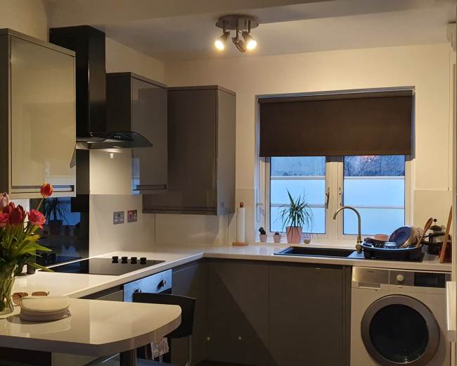 A modern kitchen with gray cabinets, a dark countertop, a window with a closed blind, and a washing machine beside the sink.