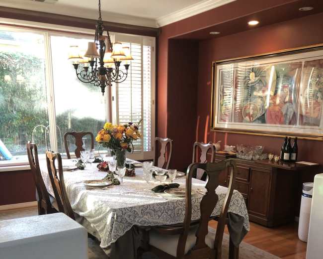 A dining room features a wooden table set with dishes and a floral centerpiece, surrounded by chairs and illuminated by a chandelier.