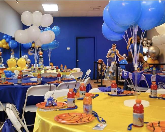The image shows a basketball-themed party setup with tables decorated in blue and yellow, featuring themed plates, Gatorade bottles, and balloons.