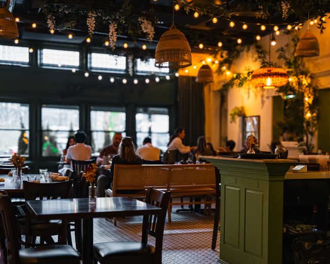 The image shows a restaurant interior with wooden tables, string lights, and greenery, filled with patrons seated at various tables.