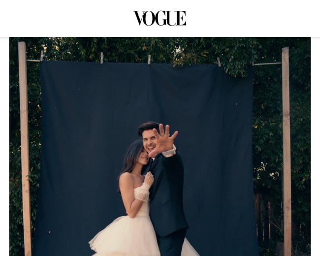 A couple poses in front of a dark backdrop, with the bride wearing a tiered wedding gown and the groom in a black suit, both smiling joyfully.