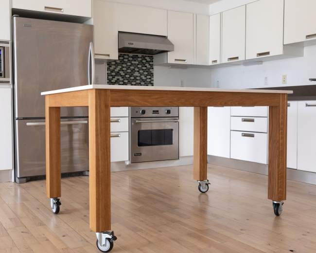 The image shows a wooden kitchen table on wheels positioned in a modern kitchen with stainless steel appliances and white cabinets.