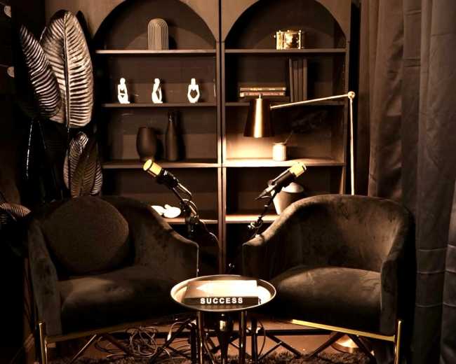 The image shows a cozy corner with two plush chairs, a small circular table labeled "SUCCESS," and a stylish bookshelf, all set against dark walls and draped curtains.