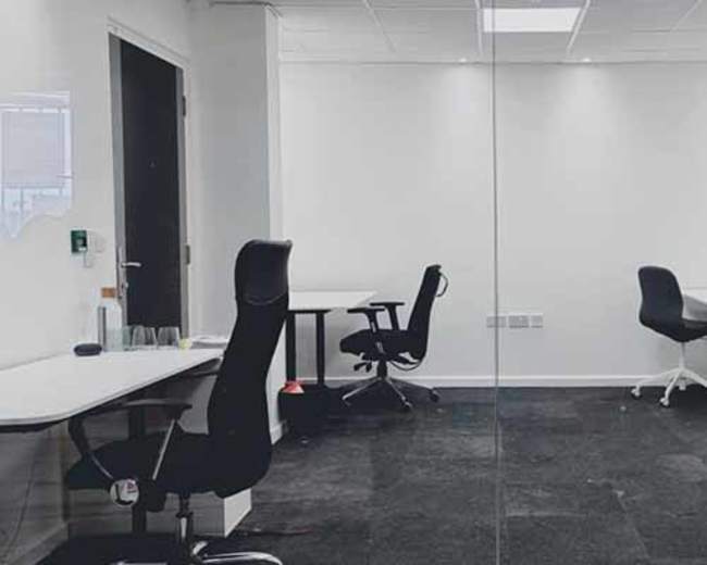 A modern office space with two desks and ergonomic chairs, separated by a glass wall.