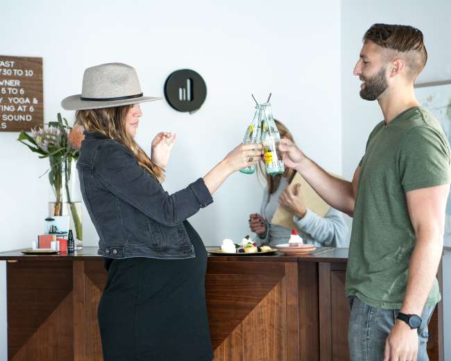A woman in a black outfit and hat clinks glasses with a man in a green shirt at a wooden bar, while another person stands in the background.