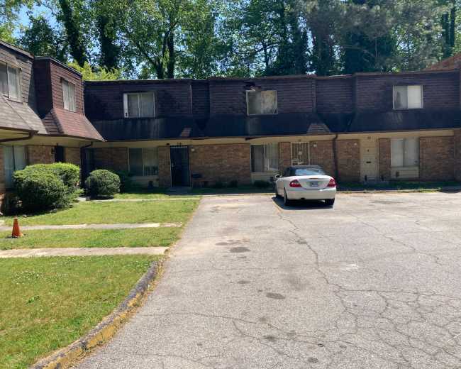 A multi-unit residential building with a gravel parking area, featuring a white car parked in front.