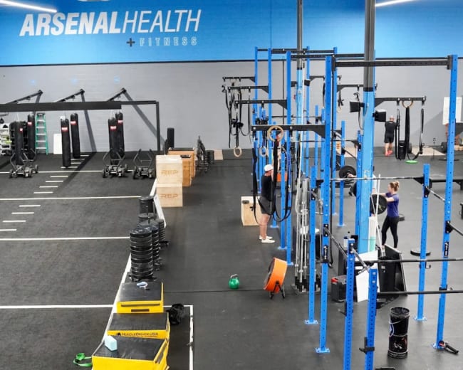 The image shows a spacious gym interior with various exercise equipment, including weightlifting stations, cardio machines, and fitness accessories, all set against a blue wall with the words "Arsenal Health + Fitness."