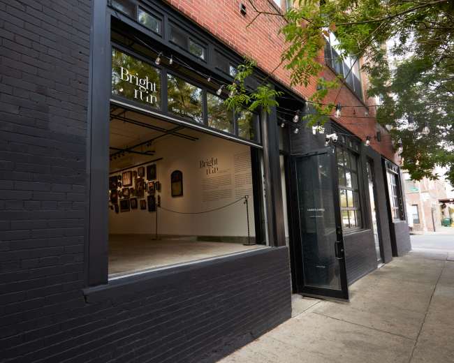 The image shows the exterior of an art gallery with large windows revealing an interior space filled with framed artworks.