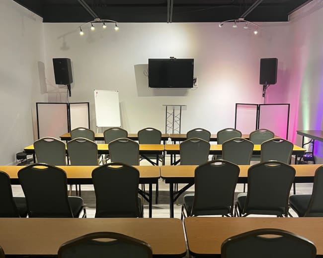The image shows a conference room setup with rows of chairs and tables facing a wall-mounted television, flanked by speakers and a whiteboard.