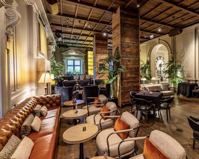 The image shows a stylish lounge area featuring a mix of brown leather seating, wooden columns, and potted plants, with tables arranged for patrons.