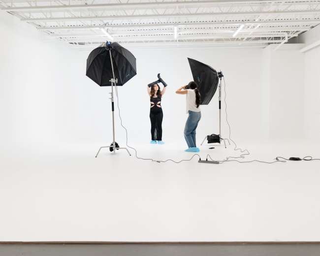 A model poses under studio lights while a photographer adjusts lighting equipment in a white backdrop studio.