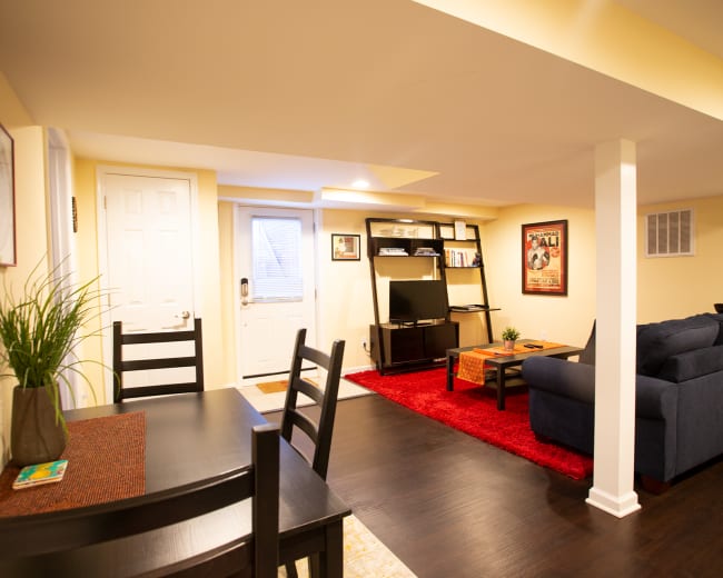 The image shows a well-lit basement living area with a dining table, a sofa, a media shelf, and a door leading outside.