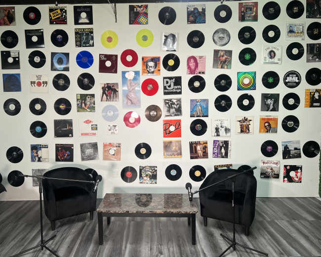 The image features a room with two black chairs and a marble table, surrounded by a wall decorated with various vinyl records.