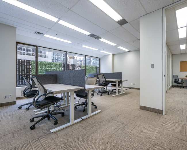 The image shows a modern office space with several desks and ergonomic chairs arranged in a well-lit area, featuring large windows and a small meeting room in the background.