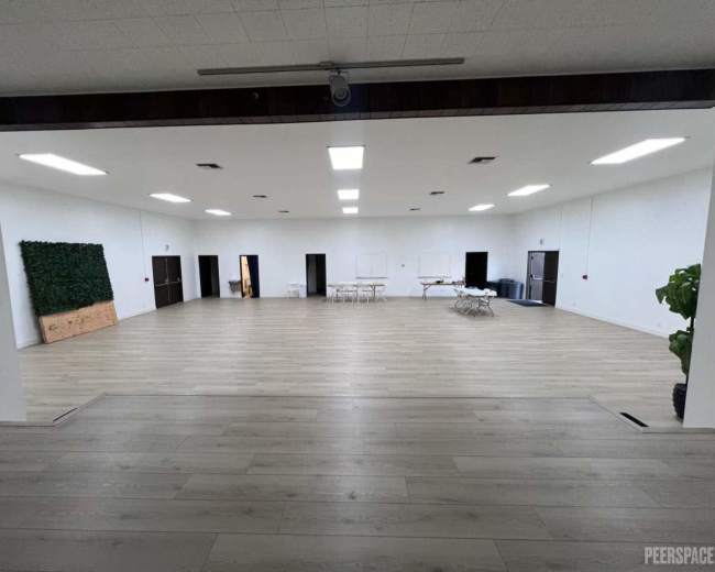 The image shows a spacious, empty interior room with wooden flooring, bright overhead lights, and simple furnishings along the walls.
