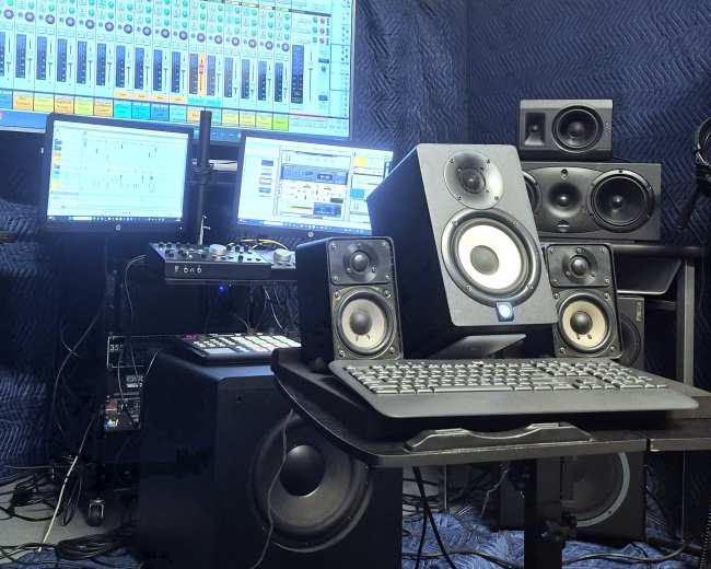 The image shows a music production setup featuring multiple speakers, a computer with two monitors, a MIDI controller, and various audio equipment arranged on a desk.
