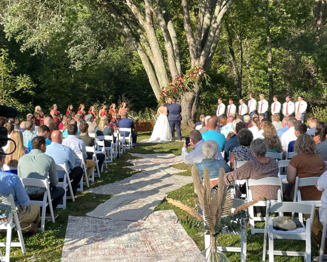 A wedding ceremony takes place outdoors under a tree, with guests seated on white chairs and a decorated aisle leading to the couple at the altar.