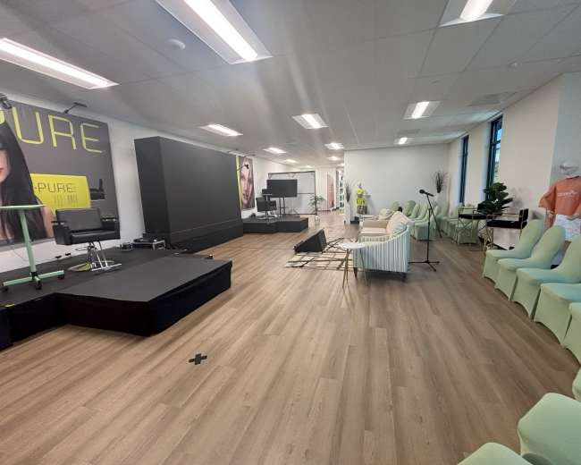 The image shows a spacious room designed for events, featuring green chairs arranged in rows, a stage area with a backdrop, and various setups including a couch and screens.