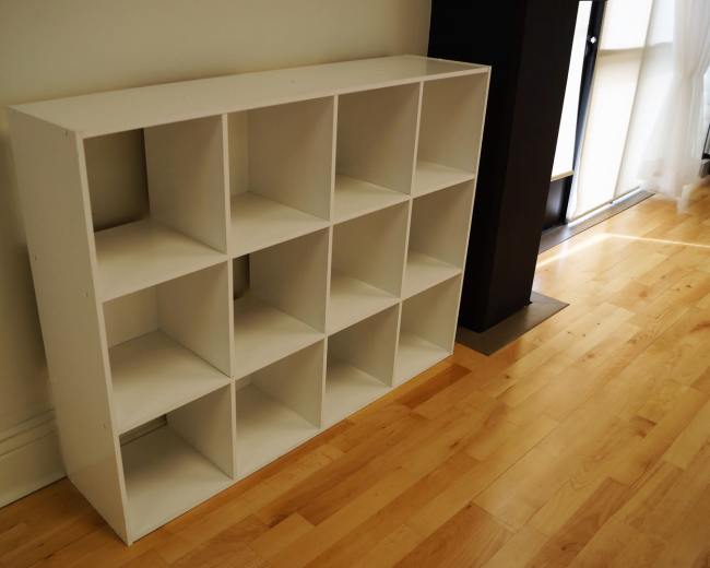 A white shelving unit with twelve square compartments positioned against a wall on a hardwood floor.