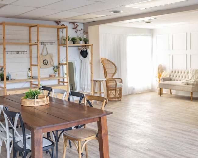 The image shows a light-filled interior space featuring a wooden dining table with chairs, shelves displaying decorative items, and a cozy seating area with a couch and a rattan chair.