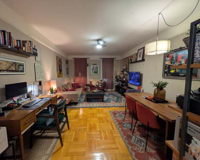 The image shows a cozy living room and workspace area furnished with a sofa, two desks with computers, and various decorative elements, including artwork and plants.