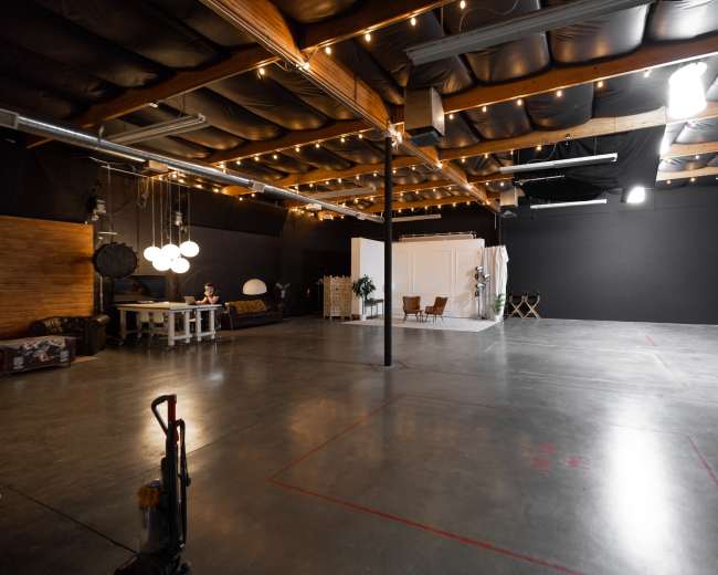 The image shows a spacious industrial studio with concrete flooring, exposed beams, and various lighting fixtures, including string lights overhead.