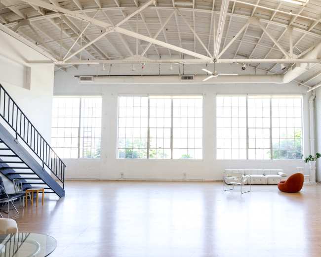 The image shows a spacious, well-lit interior with large windows, a staircase, a white sofa, and a singular orange chair.